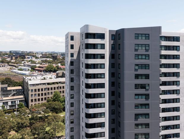 Exterior view of a contemporary multi-storey apartment building in an urban setting. The building features dark grey and light grey facade panels, repetitive rectangular windows, projecting horizontal balconies, and a flat roofline. Other urban buildings and tree-lined streets are visible in the background.