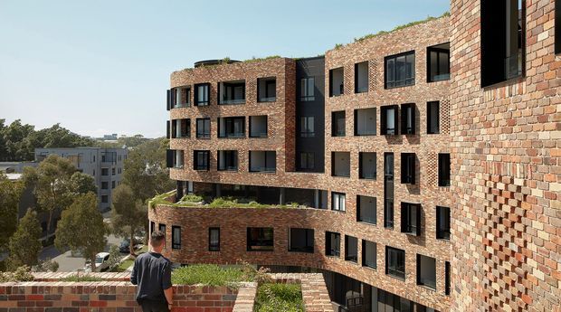 Exterior architectural view of a multi-storey contemporary brick apartment building with a curved facade, extensive black-framed windows, recessed openings, and planted rooftop and balcony terraces. The image is taken from an elevated terrace or roof, with surrounding urban greenery and neighbouring buildings visible in the background.