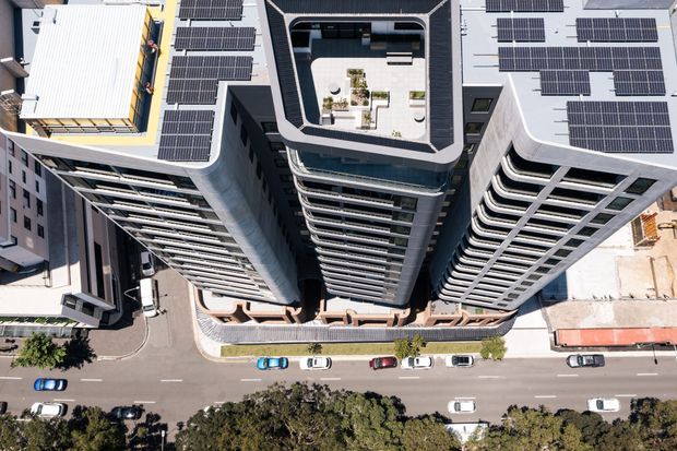 Aerial top-down view of a dense multi-storey urban apartment or commercial complex with several curved and angular towers. The flat rooftops are covered with extensive solar panel arrays, and one central rooftop contains a landscaped communal terrace with seating and planting. Streets, parked cars, and mature trees surround the development.