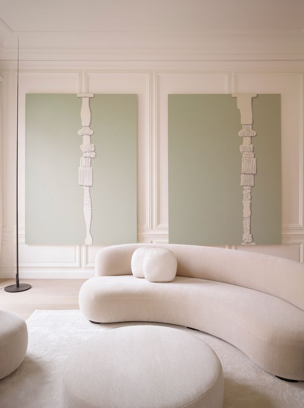 A softly lit, refined living room with a large curved cream boucle-style sofa and matching rounded ottoman on a pale rug. The walls feature elegant decorative moulding and two oversized muted sage-green artworks with sculptural white vertical motifs. A slim black floor lamp stands at the left, creating a calm, monochromatic interior with subtle classic detailing.