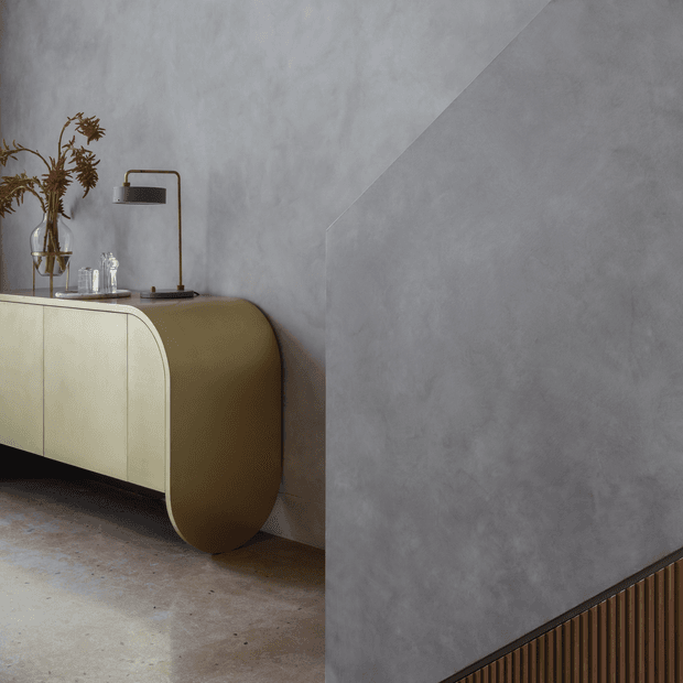 A refined contemporary interior vignette featuring a curved olive-beige sideboard against a textured grey plaster wall. The sideboard has rounded ends and pale cabinet fronts, with a slim brass-toned table lamp, glassware, and dried botanicals arranged on top. A vertical timber slat feature appears along the lower right wall, and the floor has a raw concrete-like finish.