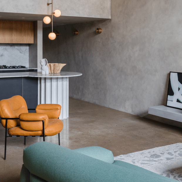 A refined open-plan living room and kitchen with a raw, textured grey plaster wall, polished concrete flooring, dark kitchen cabinetry, and a sculptural rounded island with a fluted cylindrical base. A caramel leather lounge chair, teal upholstered sofa, spherical pendant lights, and a minimalist wall-mounted television create a warm contemporary interior with sculptural mid-century influences.
