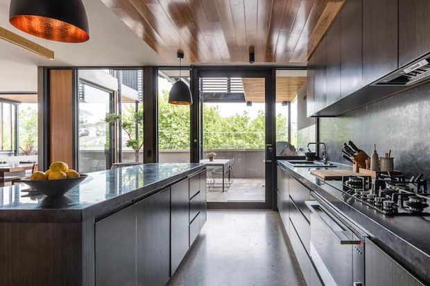 A sophisticated contemporary galley kitchen with a large central island, dark fixed cabinetry, stone benchtops, integrated appliances and a gas cooktop. Black-framed glazed doors open to a bright outdoor terrace, while a warm timber-lined ceiling and pendant lights add contrast to the dark joinery and polished concrete-like floor.