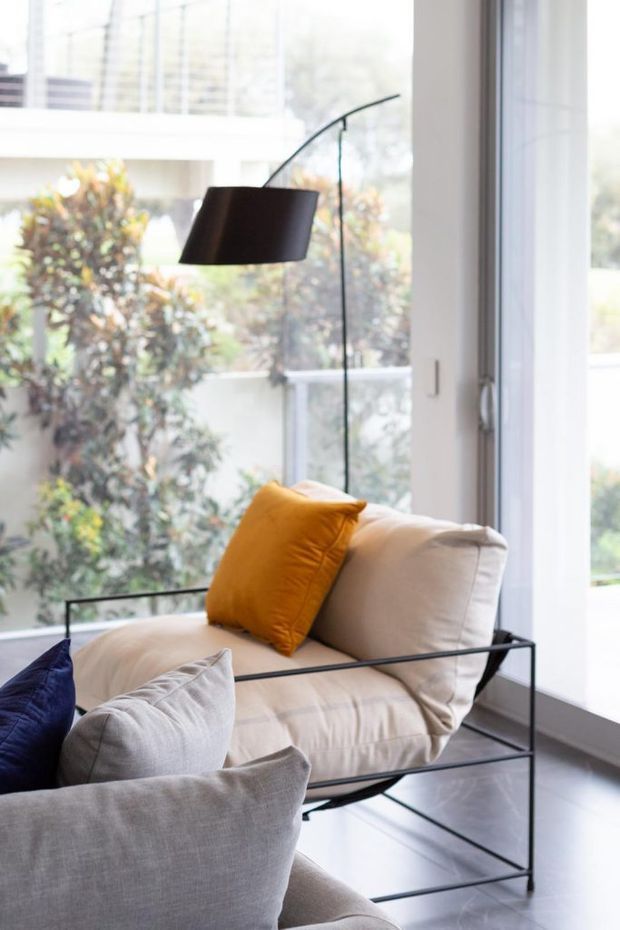 Bright contemporary living room corner featuring a light upholstered lounge chair with black metal frame, layered grey, cream and mustard cushions, and a tall arched floor lamp. Floor-to-ceiling glazing provides abundant natural light and views to greenery outside. The room has a polished dark floor and a calm, refined neutral palette with a mustard accent.