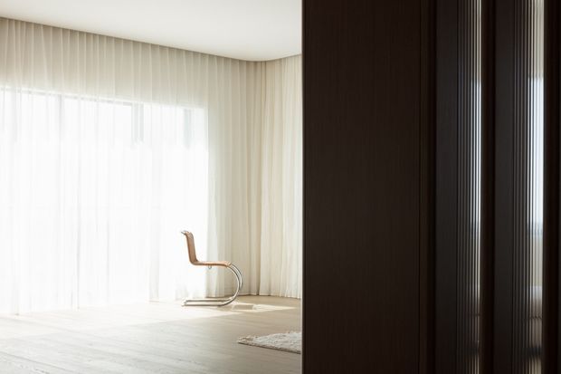 Bright, minimalist interior viewed past a dark timber partition or doorway. The room features floor-to-ceiling sheer curtains, pale timber flooring, a sculptural wooden chair, and abundant diffuse natural light. Dark vertical timber detailing creates a strong contrast at the right side of the image.