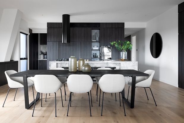 Open-plan contemporary kitchen and dining area with a long dark dining table surrounded by white upholstered chairs. The kitchen features dark timber-look full-height cabinetry, a pale stone island benchtop, integrated appliances, and a black cylindrical rangehood. Light timber flooring, white walls, large glazing, and a round black wall mirror create a clean, high-contrast interior.