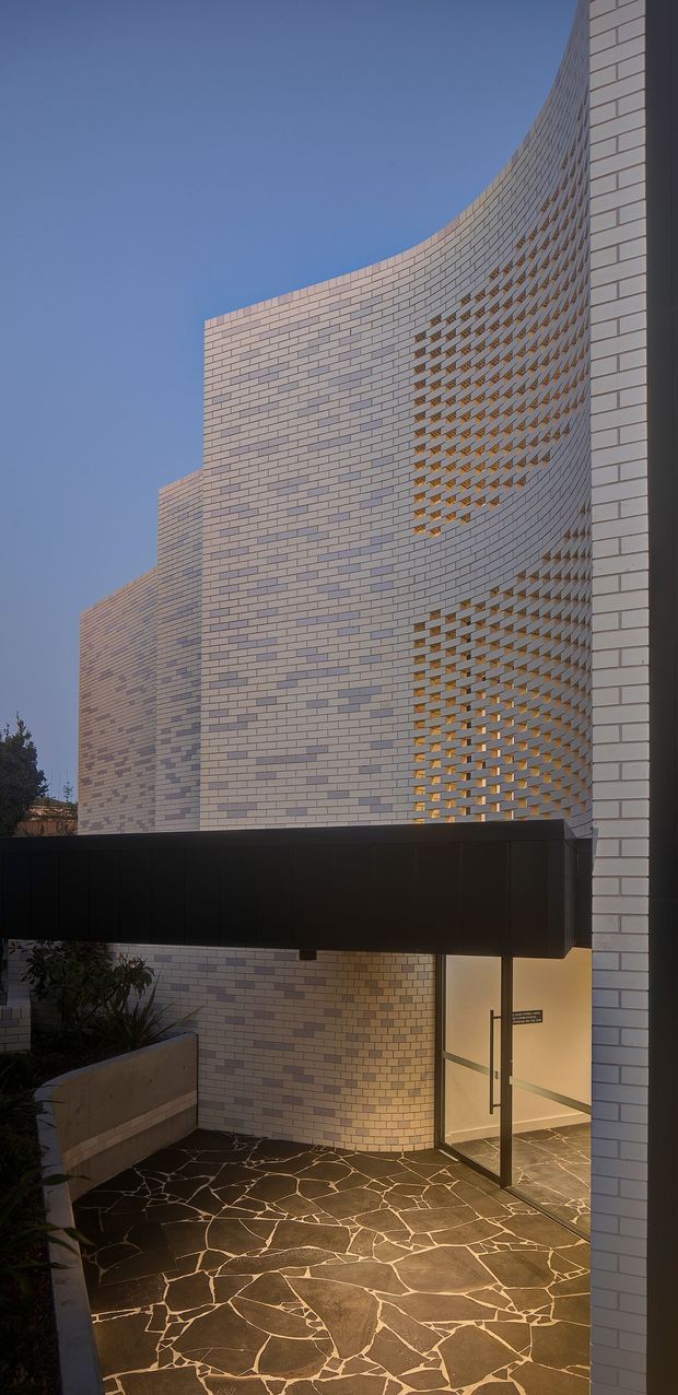 Exterior architectural view of a contemporary building with sweeping curved walls clad in light-coloured brick. A perforated brick screen creates a patterned facade with warm interior light visible behind it. The entrance is sheltered by a projecting dark metal canopy, with irregular natural stone paving and planted raised beds in the foreground.