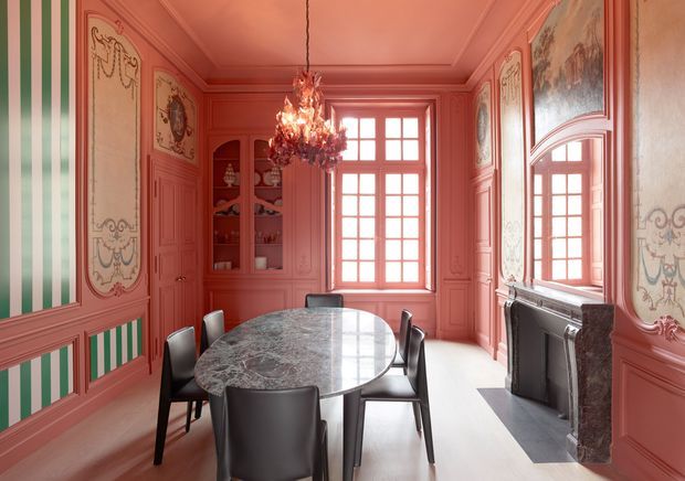 A richly detailed formal dining room in coral-pink tones, featuring ornate decorative wall panelling, painted mouldings, striped green-and-white inset panels, a large marble oval dining table with dark contemporary chairs, a crystal chandelier, tall French doors or windows, and an elaborate fireplace with an overmantel mirror. The space combines historic decorative architecture with modern furniture.