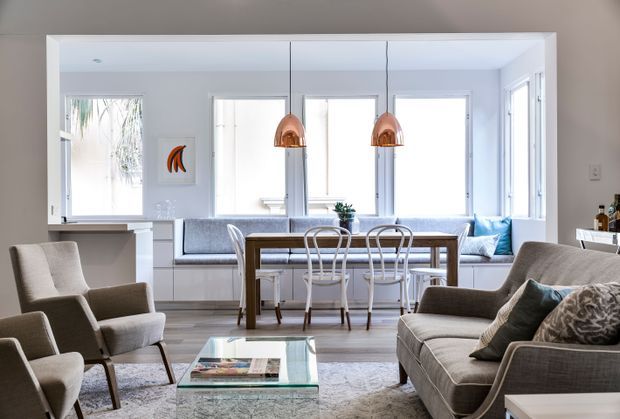 Bright open-plan living and dining area with a neutral palette, pale timber flooring, large windows, and a white built-in kitchen or service counter at the rear. The room contains a grey upholstered sofa and armchairs, a timber dining table with metal-frame chairs, a glass coffee table, and two copper pendant lights. The space feels airy, refined, and comfortably contemporary, with abundant natural light.