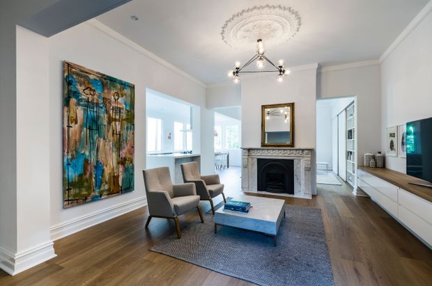 Bright, refined living room with dark timber flooring, pale walls and ceiling, ornate plaster ceiling medallion, decorative fireplace mantel, framed artwork, two upholstered lounge chairs, a low square coffee table, and streamlined white media cabinetry. Open doorways connect to adjacent light-filled spaces, while a large abstract painting provides a strong colour accent.