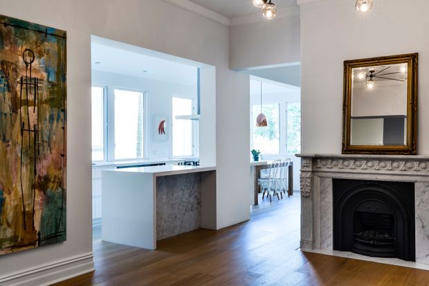 A bright, refined interior view from a living room toward an open white kitchen and dining area. The room features warm timber flooring, pale walls, a sculptural ornate fireplace with a dark firebox, a framed mirror above the mantel, decorative wall artwork, and contemporary pendant lighting. The kitchen includes a white island with a grey veined stone face, while the dining area is visible beyond.