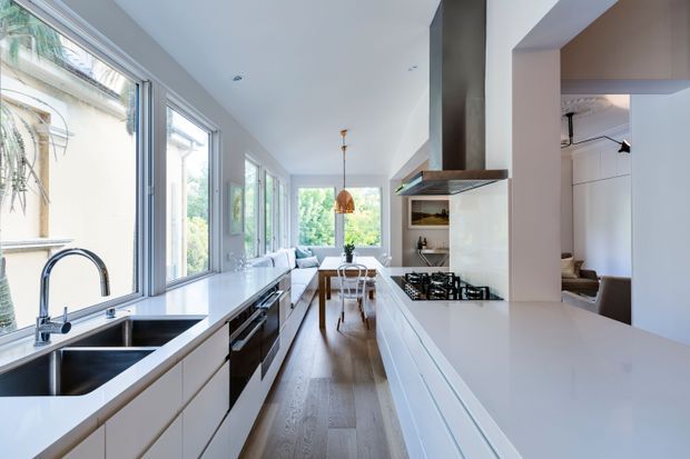 Bright, elongated contemporary kitchen with a streamlined white island and cabinetry, double stainless-steel sink beneath expansive windows, integrated appliances, gas cooktop, and a prominent dark metal range hood. Warm timber flooring runs through the space toward a compact dining area and glazed garden outlook. The room is filled with natural light and has a clean, refined open-plan feel.
