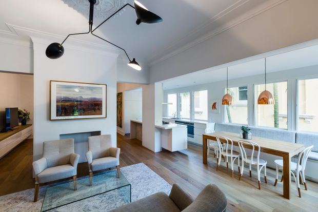 Open-plan contemporary living, dining and kitchen space with white walls, decorative ceiling cornices, timber flooring, a glass coffee table, upholstered armchairs, a dining table with white chairs, and a bright kitchen zone beyond. Black sculptural pendant lighting and copper-toned dining pendants add contrast, while large windows provide abundant natural light.