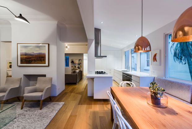 Open-plan contemporary home interior combining a living room, kitchen and dining area. The space features white cabinetry and walls, warm timber flooring, a central kitchen island, integrated stainless-steel appliances, a large timber dining table, upholstered dining chairs, pendant lighting, and a compact seating area with framed artwork and armchairs. Abundant glazing brings in natural light, while copper-toned pendant shades provide a warm accent.
