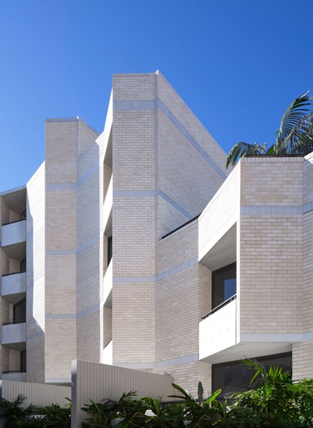 Exterior architectural photograph of a multi-storey contemporary residential building with strong geometric massing, pale brick cladding, projecting vertical brick volumes, recessed balconies, and crisp white structural elements. Tropical palms and landscaping appear in the foreground against a clear blue sky.