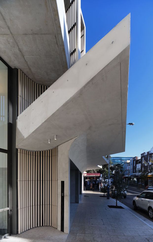 Street-level view of a contemporary urban building facade with prominent cantilevered raw concrete balconies and soffits, vertical dark and light screening fins, large glazing, and a recessed entrance. The building sits directly along a paved commercial streetscape with neighbouring multi-storey buildings visible in the background under a clear blue sky.