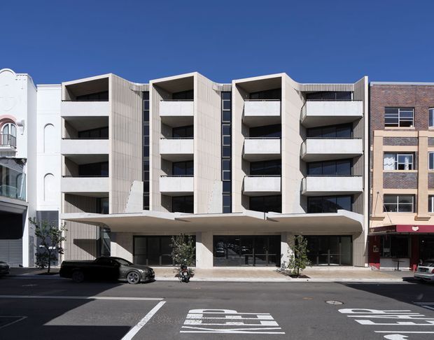 Street-facing view of a contemporary multi-storey apartment building with a pale concrete and vertically ribbed facade, repeated recessed balconies, dark vertical glazing strips, and a broad projecting entry canopy. The building sits within an urban streetscape between older neighbouring commercial or residential buildings, with marked roadway in the foreground.