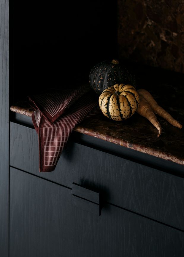A moody close-up of dark kitchen cabinetry with a projecting rectangular pull, topped by a dark mottled stone benchtop. Decorative gourds and carrots sit on the counter beside a draped burgundy patterned textile. The scene is styled with near-black cabinetry, warm earthy accents, and low dramatic lighting.