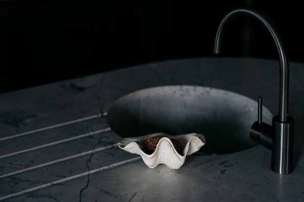A dark, moody close-up of a sculptural black stone or charcoal-grey bathroom basin with a tall curved black tap. The basin sits on a veined dark stone benchtop, with a pale shell-shaped decorative vessel containing a small amount of organic material in front. Minimal black background and dramatic directional lighting emphasise the tap and basin forms.