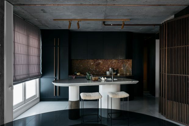 A dark, refined contemporary kitchen centred on a slim island with two upholstered stools. Matte near-black cabinetry, a dark stone island benchtop, and a dramatic brown-toned stone splashback contrast with pale flooring and a concrete ceiling. Full-height glazing with a Roman blind is visible on the left, while a vertical timber slat screen frames the right side. Minimal timber track lighting and restrained styling create a moody, luxurious atmosphere.