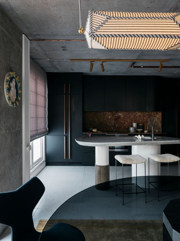 A dramatic, moody contemporary kitchen with a large rounded island, dark built-in cabinetry, a richly veined brown stone splashback, and integrated sink and tapware. The island has white cylindrical supports with dark timber or stone bases and is paired with upholstered bar stools. Concrete-look walls and ceiling, pale large-format flooring, a dark curved floor zone, a sculptural pendant light, track lighting, and a pink Roman blind create a refined, high-contrast interior.