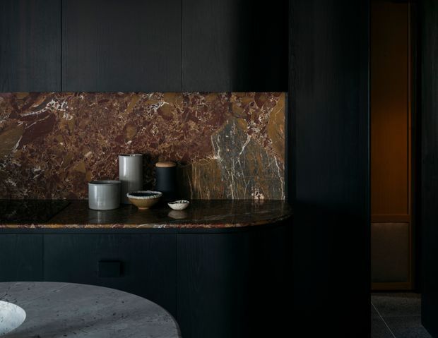 Moody close-up of a dark, sophisticated kitchen or bar-like built-in joinery. Black or charcoal cabinetry frames a richly veined brown and burgundy stone splashback and matching dark stone counter, with ceramic vessels arranged on the bench. A rounded grey stone or concrete island edge is visible in the foreground, and a dimly lit adjoining passage or cabinetry appears at right.