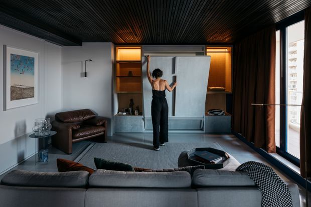 A sophisticated compact living room with a large grey sectional sofa in the foreground, a dark leather armchair, grey rug, and a low black coffee table. The rear wall features a concealed wall-mounted television or display behind a large sliding panel, integrated warm-lit timber shelving and cabinetry, and a small bar or display niche. Dark ribbed timber ceiling, full-height dark curtains, large windows, and grey painted walls create a refined, moody contemporary interior.