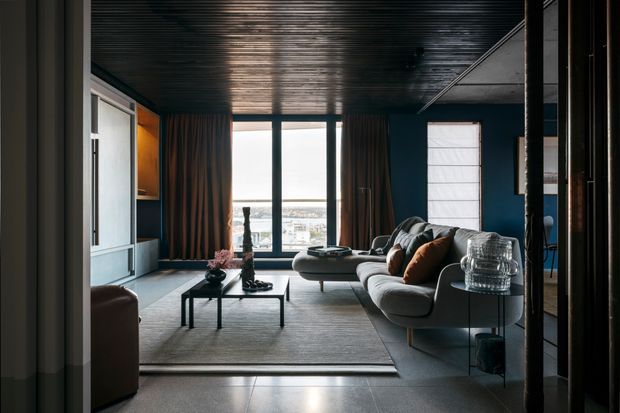 A moody contemporary living room with a large grey sectional sofa, dark blue walls, full-height glazed doors opening to a distant city or water view, and floor-to-ceiling curtains. The room features a dark ribbed ceiling, pale flooring, a large neutral rug, black coffee tables, built-in cabinetry, and warm timber-toned joinery. The composition is framed from an adjoining circulation space, with a reflective glass or dark-framed opening at the right.