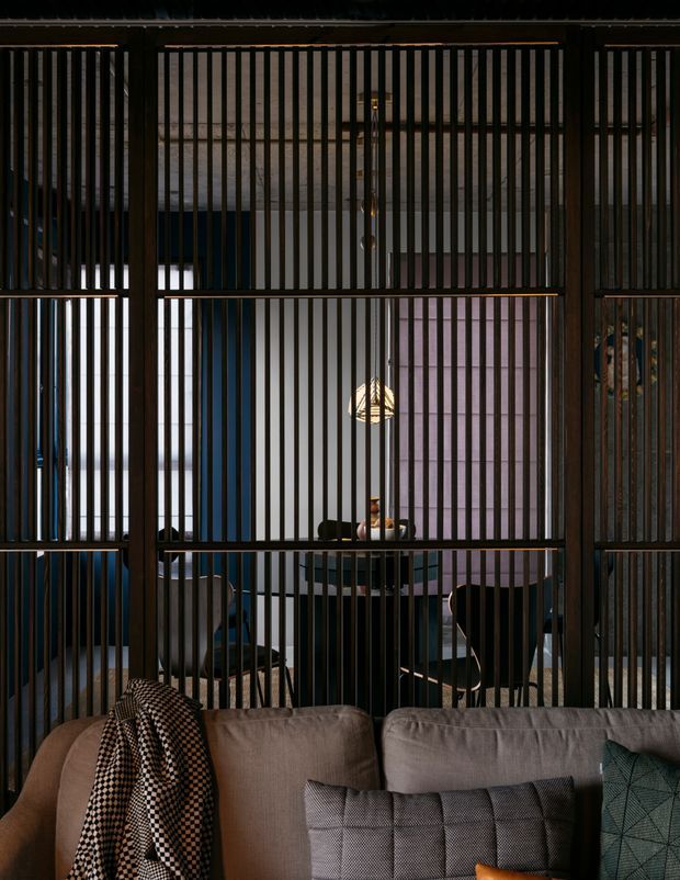 Moody interior view from a living room toward a dining area, screened by a full-height dark timber slatted partition. A sofa with patterned cushions and a throw occupies the foreground, while a round dining table, chairs and pendant light are visible behind the partition. The space uses dark vertical timber detailing, deep blue and mauve wall tones, and subdued warm lighting.