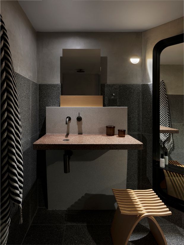 A compact, moody bathroom with dark textured wall and floor tiles, a terrazzo-style floating vanity with integrated backsplash, wall-mounted basin mixer, and a large black-framed arched mirror. A sculptural timber slatted stool sits in the foreground, with a striped textile visible at the left and in the mirror reflection. Warm wall lighting and dark finishes create a refined, contemporary atmosphere.