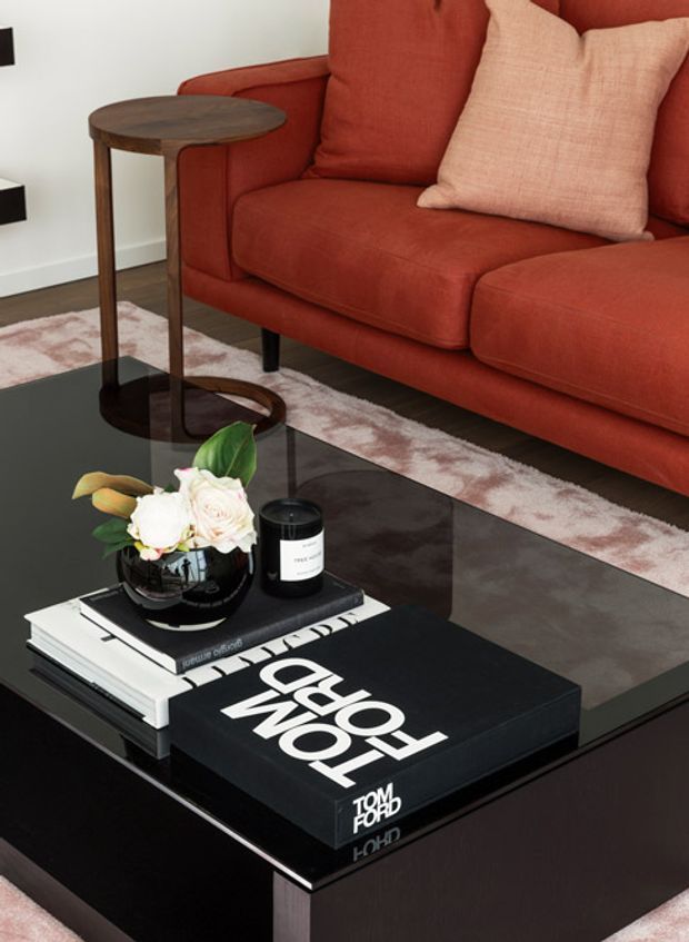 Close-up interior vignette of a contemporary living room, featuring a deep rust-red upholstered sectional sofa with a blush cushion, a dark glass coffee table styled with books, a black candle, and a floral arrangement, plus a slim round timber side table. A pale patterned rug and white wall provide contrast.
