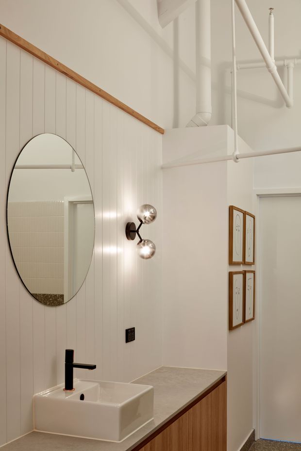A compact, warmly lit bathroom or powder room with a white vessel basin, matte black wall-mounted tap, pale vertical wall panelling, and a timber-edged stone-look vanity. An oval mirror, sculptural polished-metal wall sconce, exposed white services, framed artwork, and a partially visible tiled shower area add character. The room has a refined industrial feel softened by timber detailing and warm neutral finishes.