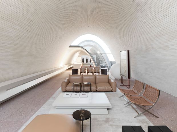 A bright, highly sculptural contemporary lounge with a long barrel-vaulted white interior and fine linear wall and ceiling detailing. A large arched glazed opening forms a dramatic focal point at the rear, while a tan leather sofa, low white layered coffee table, black side table, woven sling chairs, and a curved tan ottoman furnish the space. The room has a refined gallery-like atmosphere with strong geometric forms and abundant diffuse daylight.