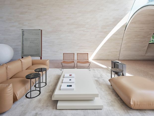 A spacious, sculptural living room with a dramatic curved, ribbed pale-wood ceiling and wall treatment. The room features low, oversized cream leather sofas, a large white layered coffee table, black slender side tables, woven lounge chairs, a pale textured rug, and an abstract artwork. Strong linear daylight enters through a curved opening, creating a refined, gallery-like atmosphere.
