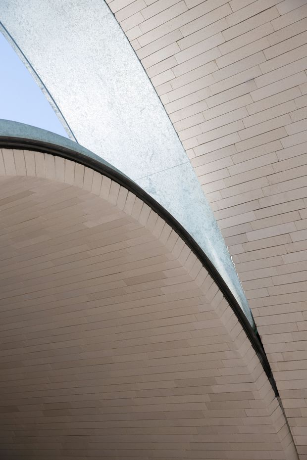 Close-up architectural view looking upward at a curved, barrel-like brick arch or soffit intersecting with a tall pale brick wall. The masonry is light beige with dark curved metal edging, while a blue-grey textured structural or cladding surface cuts diagonally across the composition. A small area of blue sky is visible at the edge. The image emphasizes geometric curves, brickwork, and structural detail rather than a complete room or facade.