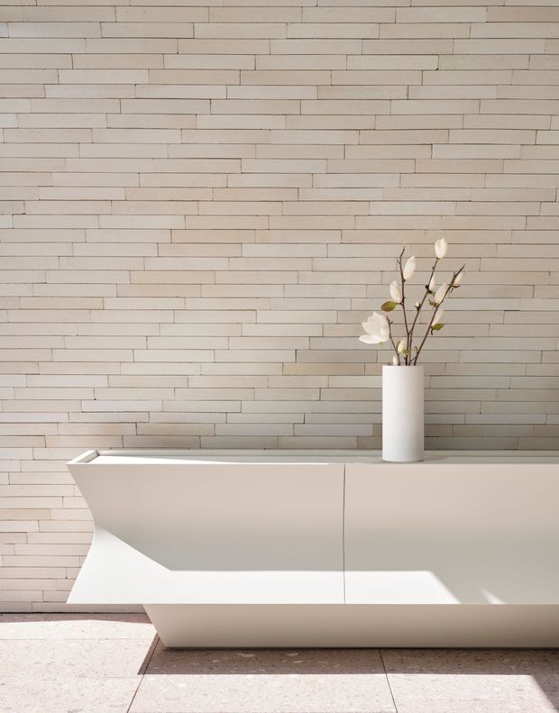 A minimalist bathroom vignette featuring a sculptural freestanding white bathtub against a full-height wall of narrow horizontal cream-beige tiles. A simple white vase with delicate branches sits on the tub ledge, while large pale stone or porcelain floor tiles and strong sunlight create a calm, refined atmosphere.