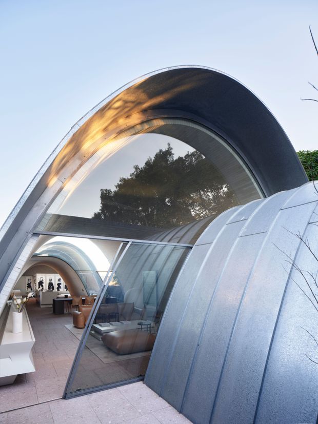 A distinctive modern building exterior with curved, barrel-vaulted forms clad in silvery metal panels. A large arched glazed opening reveals a stylish interior lounge or gallery, while the sculptural shell-like structure and sweeping roofline dominate the view. The image is photographed from outside in bright daylight, with reflections of trees in the glazing.