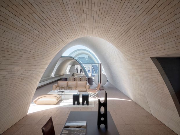 A sculptural, tunnel-like living space with a curved arched ceiling and walls lined in narrow light timber or brick-like strips. The bright minimalist room contains a low beige sofa, glass coffee table, sculptural black side tables, and a dining or work area toward the rear. An arched glazed opening brings in abundant daylight and reinforces the futuristic, organic form.