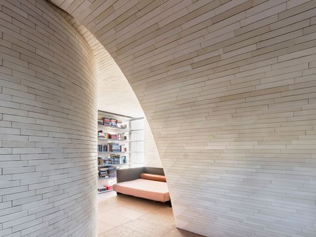 Curved architectural interior lined with narrow pale masonry blocks, forming a sculptural arched passage around a small reading nook. A low blush-pink sofa sits beside built-in open shelving filled with books, with pale tiled flooring and soft natural light. The space has a highly sculptural, minimalist character.
