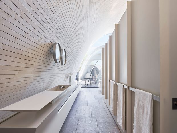 A bright, elongated contemporary bathroom with a long floating double vanity and twin round mirrors along a horizontally tiled feature wall. A freestanding bathtub sits beneath the vanity area, while a row of towel rails and pale towels lines the opposite wall. The space extends toward an arched glazed end with a small seating area, creating an airy, refined atmosphere. Pale stone-look surfaces and white finishes dominate.