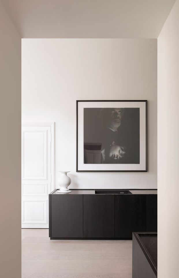 A refined minimalist interior featuring a long, dark built-in sideboard with slab-front cabinetry and a dark stone-like top. The cabinet sits against a warm white wall beneath a large black-framed monochrome artwork. A white decorative vase provides a subtle accent, while a paneled white door and pale timber floor are visible to the left. The space is softly lit, neutral, and highly uncluttered.
