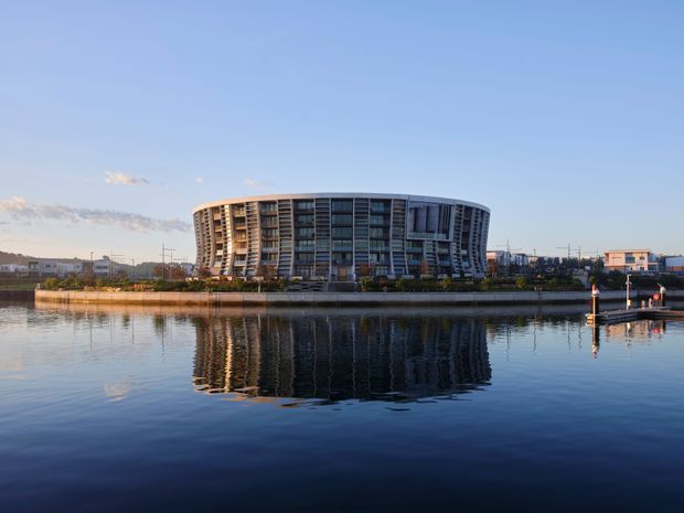 Wide exterior view of a large curved contemporary multi-storey building beside calm water, with extensive glazed façades, balconies, and a strong reflection across the waterfront. The scene includes a low urban shoreline, a small pier or dock, and clear blue sky in warm daylight.