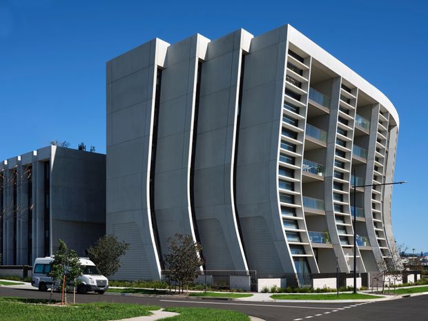 Exterior architectural photograph of a contemporary multi-storey apartment or commercial building with a sculptural curved facade. The structure uses dark grey panelled surfaces, strong vertical fins, extensive recessed balconies, and white structural framing. It is viewed from a landscaped street frontage under a clear blue sky, with a smaller adjoining building visible to the left.