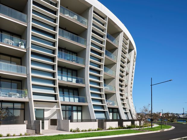 Exterior view of a contemporary multi-storey apartment building with a distinctive curved facade, repeated recessed balconies, extensive glazing, glass balustrades, and strong horizontal sunshade bands. The building fronts a landscaped verge and paved roadway under a clear blue sky.