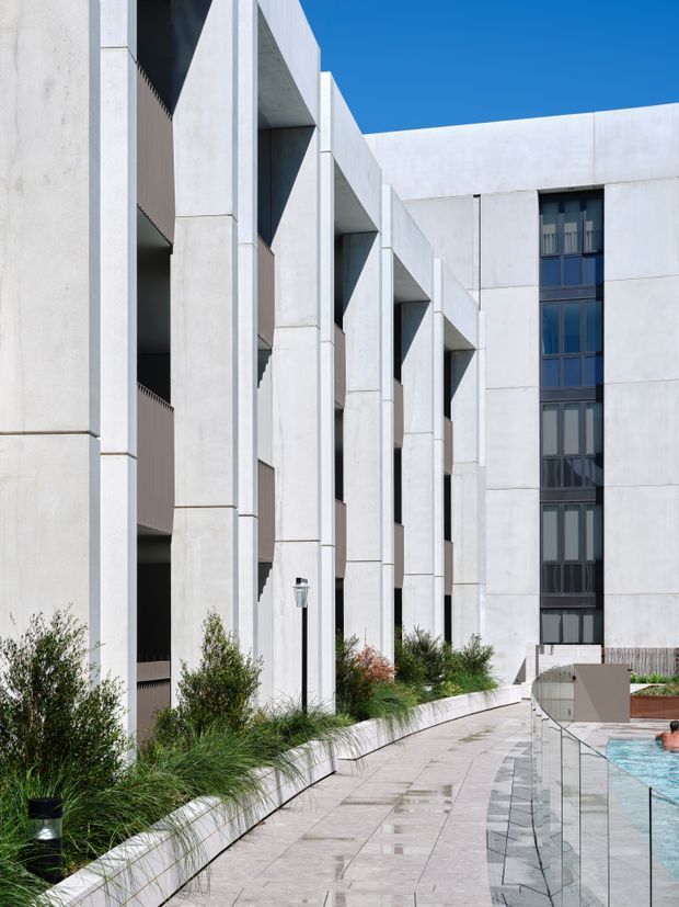 A contemporary multi-storey building courtyard with a strongly geometric concrete facade. Repeated deep vertical recesses and projecting structural frames line the left side, while dark glazed openings and a taller white wall define the far elevation. A landscaped planter with grasses and shrubs borders a pale paved walkway, which runs beside a swimming pool enclosed by a clear glass balustrade. The image is brightly lit under a clear blue sky.