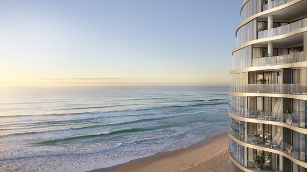 Exterior architectural view of a tall, contemporary curved apartment building directly beside a sandy beach and calm ocean. The facade features rounded projecting balconies, extensive floor-to-ceiling glazing, dark metal or glass balustrades, and pale concrete bands. The oceanfront setting dominates the scene.