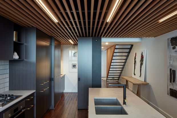 A sophisticated contemporary kitchen and circulation space with dark timber cabinetry, a pale stone island with integrated double sink, timber flooring, and a striking slatted timber ceiling with linear recessed lighting. A partially visible open-riser stair connects to the rear, while shelving, artwork, and a timber console add detail to the adjacent area.