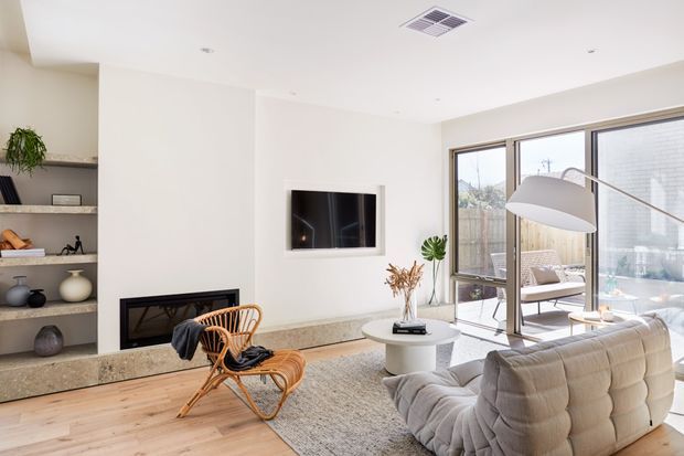 Bright, open-plan contemporary living room with a large cream modular sofa, sculptural floor lamp, round coffee table, woven rattan lounge chair, textured rug, built-in fireplace with stone surround, wall-mounted television, and full-height glazed doors opening to an outdoor seating area. Built-in shelving and neutral finishes create a calm, refined interior.