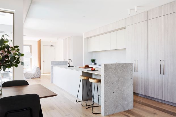 Bright, open-plan contemporary kitchen and dining area with extensive pale timber cabinetry, a white island, and a contrasting textured stone feature panel. Two bar stools sit at the island, while a dark dining table and chairs occupy the foreground. The space has white walls and ceiling, light timber flooring, minimalist detailing, and abundant natural light.