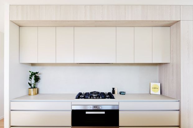 Minimalist contemporary kitchen with a symmetrical built-in cabinetry wall, pale timber-look framing and lower drawers, white upper cabinets, a light benchtop, integrated black oven, and a compact gas cooktop. The bright, uncluttered design includes a small plant and subtle accessories.
